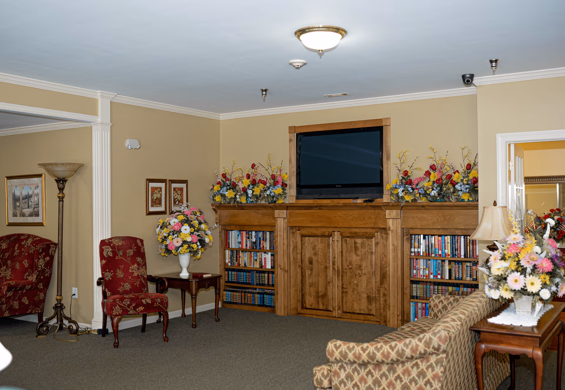 A cozy living room area with beige walls and carpeted floor. There is a wooden entertainment center with a flat-screen TV mounted above it, flanked by bookshelves filled with books and decorated with colorful flower arrangements. The room features patterned upholstered chairs and a sofa, a wooden side table with a lamp and a flower arrangement, and framed artwork on the walls.