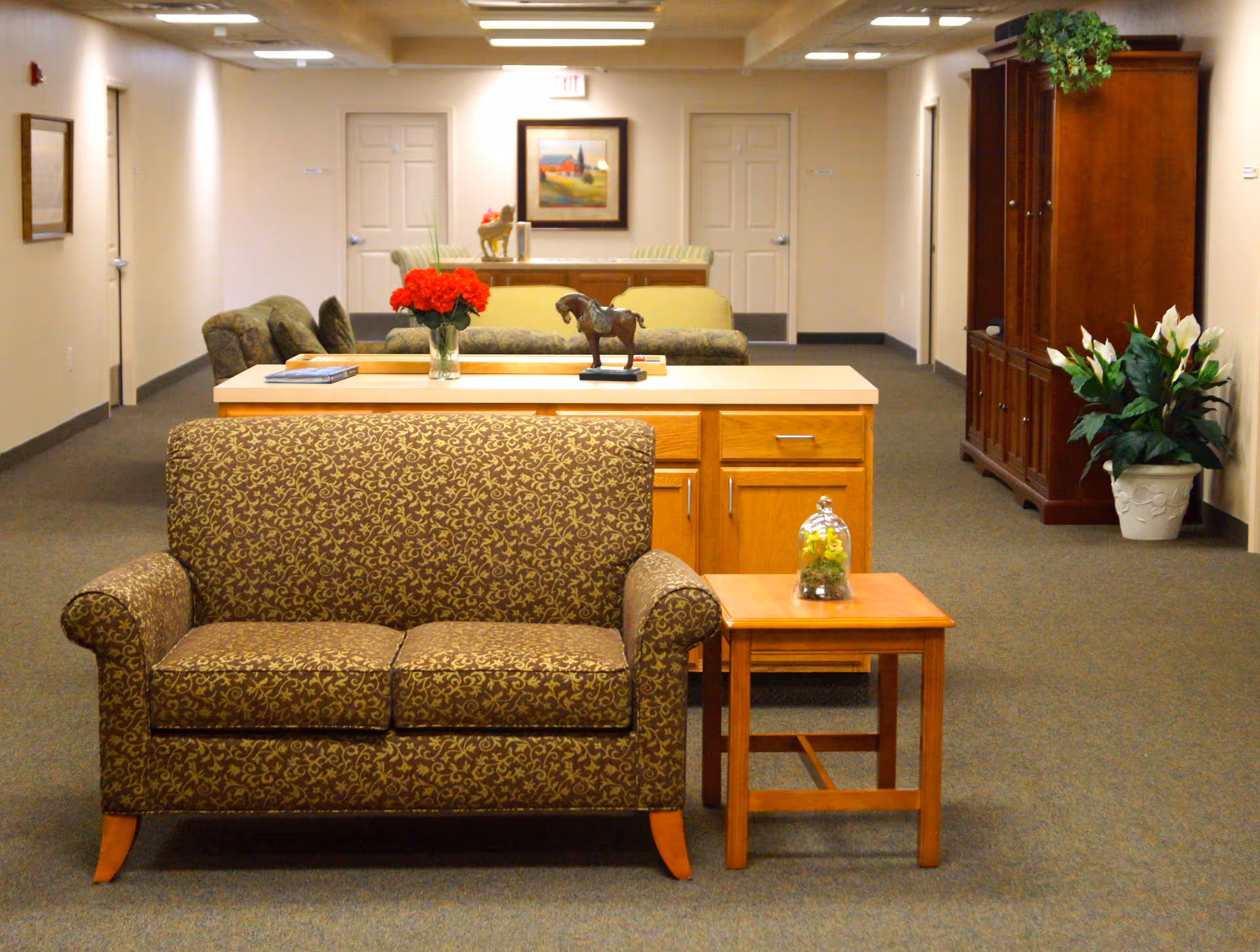 Interior view of a senior living facility lounge area with patterned brown loveseat, wooden side table with a small plant under glass, wooden cabinet with drawers, a vase of red flowers, a horse figurine, and framed artwork on the wall. The space is carpeted and has multiple doors along the walls.