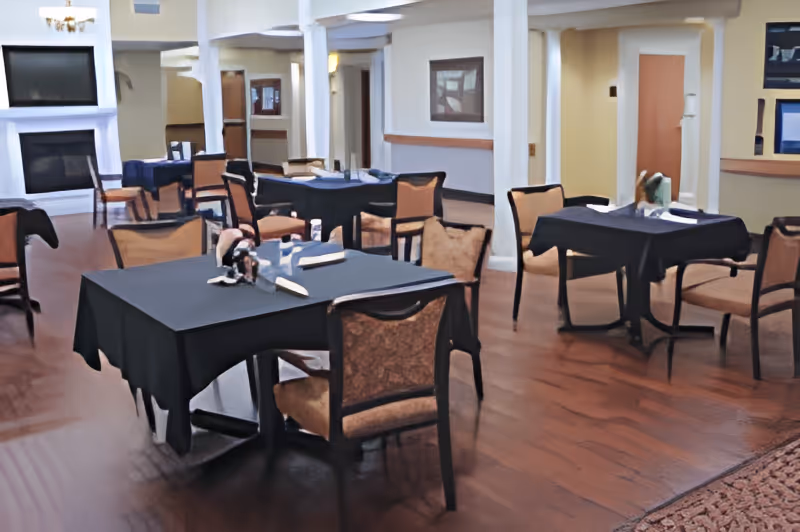 Interior view of a dining area with several tables covered in black tablecloths and cushioned chairs arranged around them. The room has wooden flooring, white walls with columns, and a fireplace with a TV mounted above it in the background.