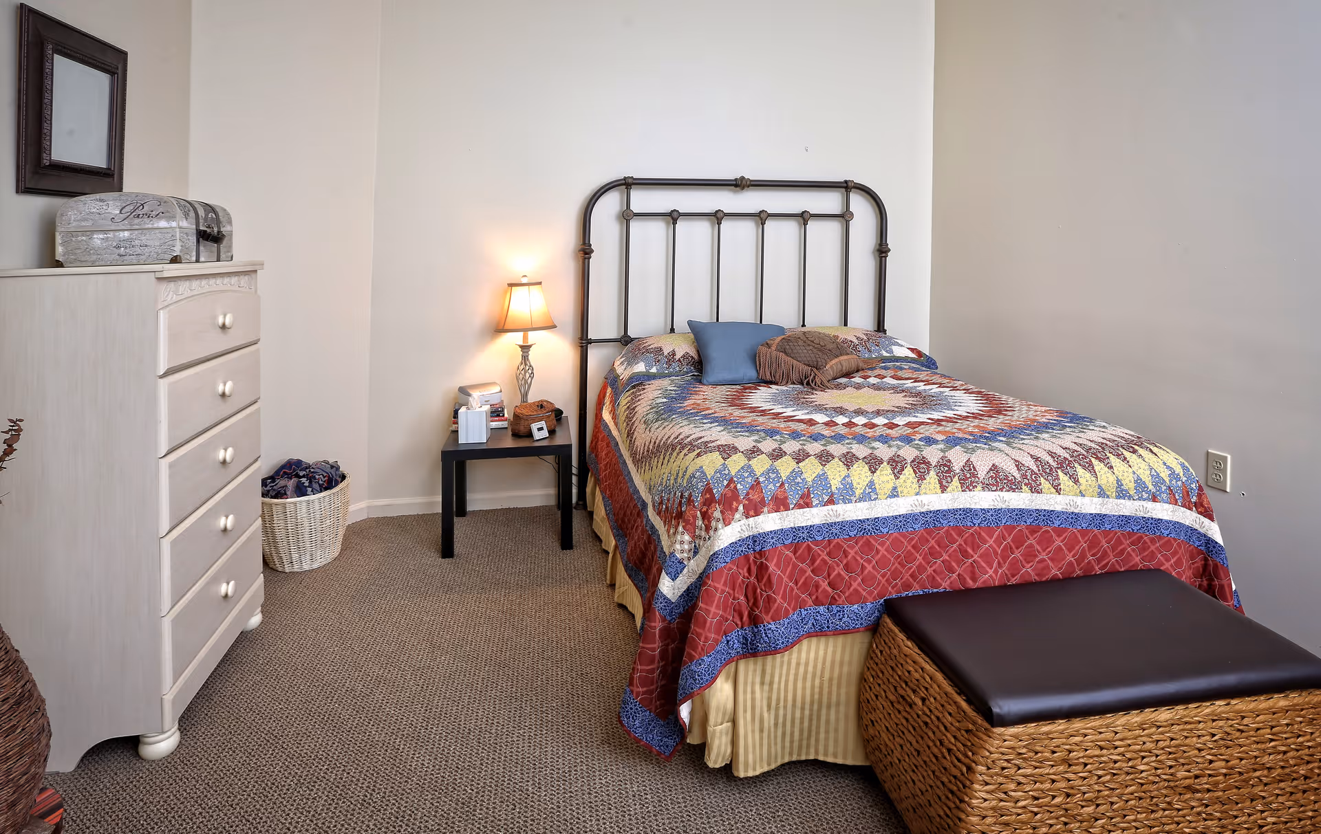 A tidy bedroom with a metal-framed bed covered by a colorful quilt, a nightstand with a lamp, a dresser, and a woven ottoman.