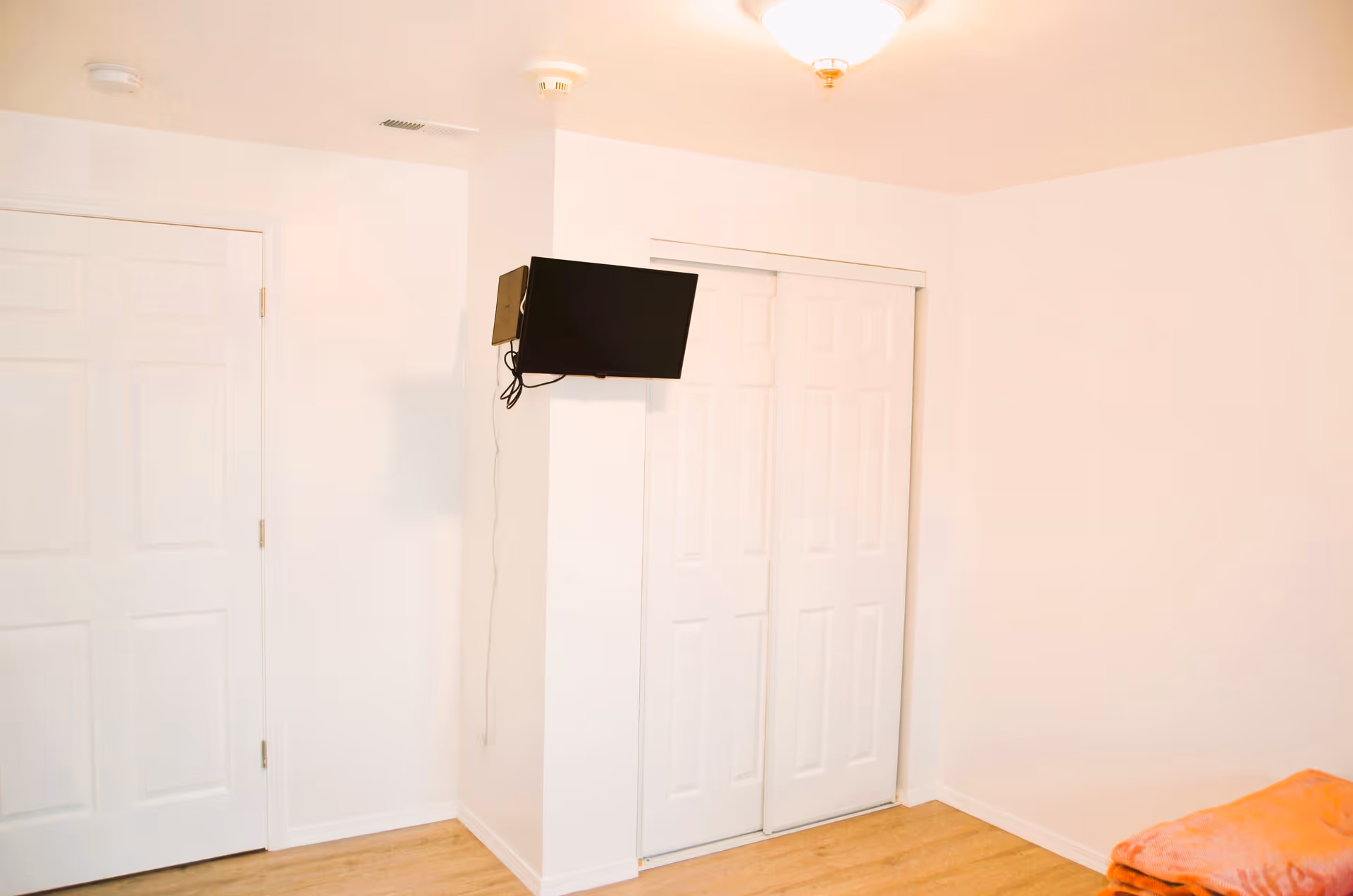 A simple room with white walls and light wood flooring. There is a closed white door on the left, a wall-mounted flat screen TV in the center, and white sliding closet doors on the right. An orange blanket is partially visible in the bottom right corner.