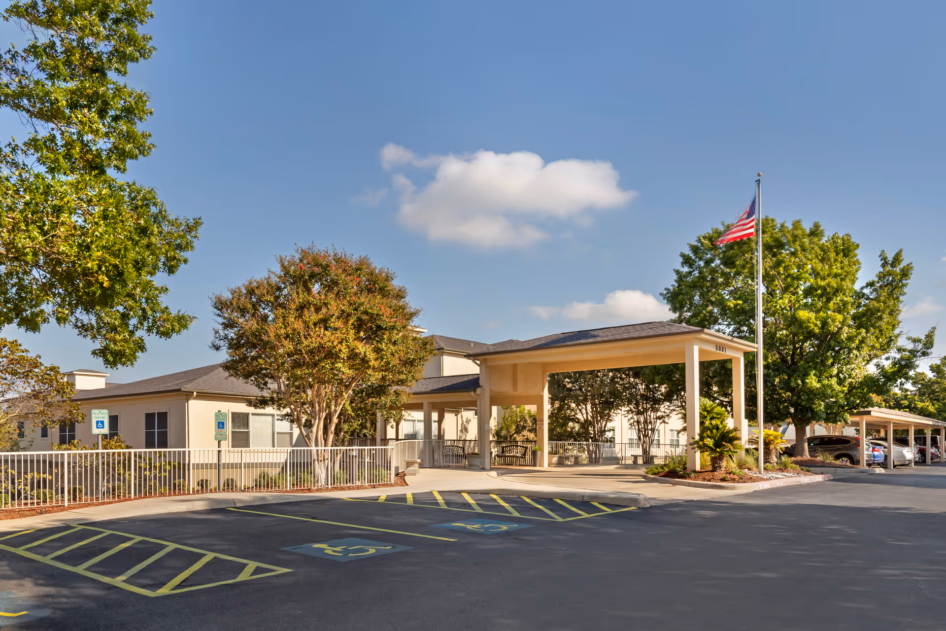 Exterior view of Brookdale Hamilton Wolfe facility showing the entrance with a covered drop-off area, an American flag on a flagpole, several trees, and a parking lot with designated handicapped parking spaces.