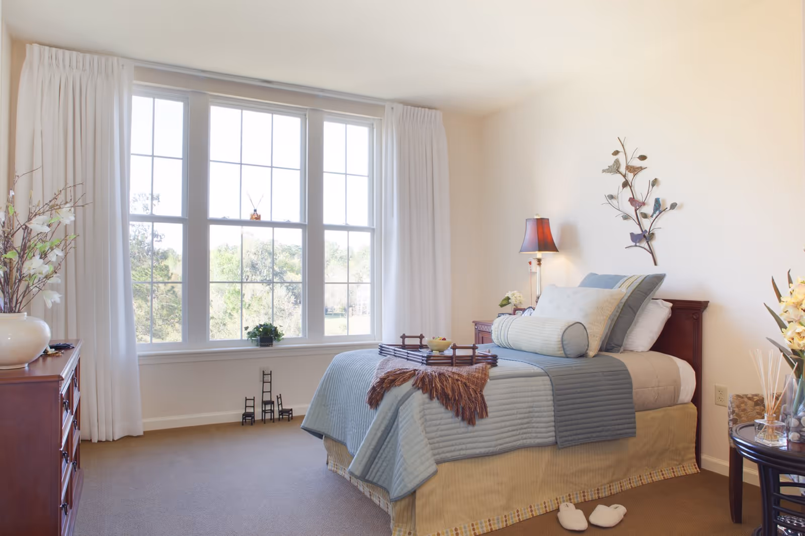 A bright and cozy bedroom with a large window letting in natural light. The bed is neatly made with blue and beige bedding, decorative pillows, and a brown throw blanket. A wooden nightstand with a lamp and flowers is beside the bed. There is a wooden dresser with a vase of flowers on the left side, and a small round table with flowers and decorative items on the right. White curtains frame the window, and a wall decoration with birds is above the bed. Slippers are placed on the carpeted floor near the bed.