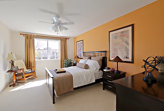 Well-lit bedroom with a bed, nightstands, dresser, ceiling fan, armchair by a window, and a warm orange accent wall.