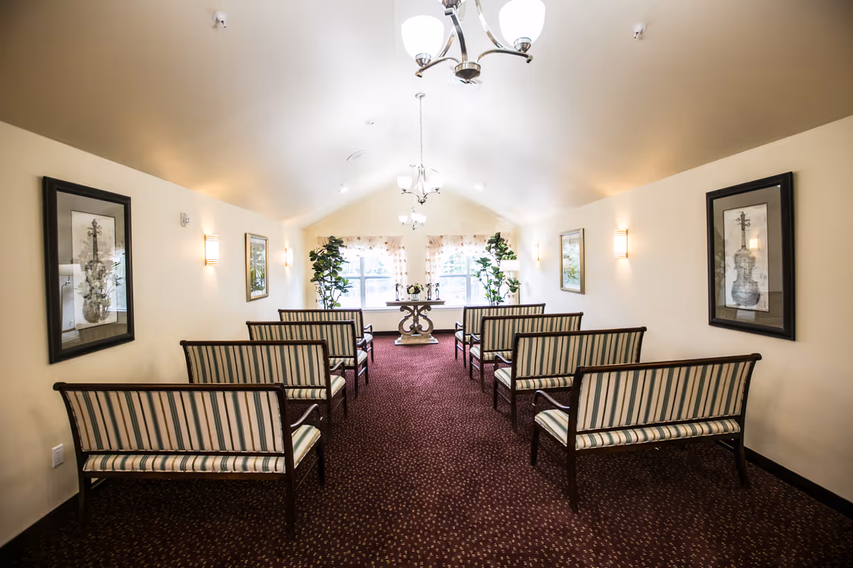 A bright, carpeted meeting room with rows of striped wooden benches facing a small table and window at the far end.