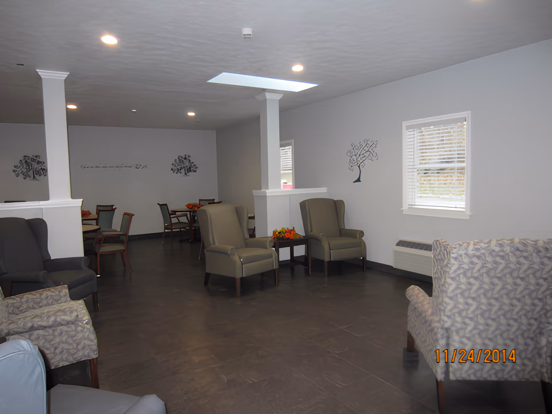 A spacious common area in a senior living facility with multiple armchairs and small tables arranged around the room. The walls are light gray with decorative tree artwork, and there is a window with blinds letting in natural light. The floor is dark, and the ceiling has recessed lighting and a skylight.