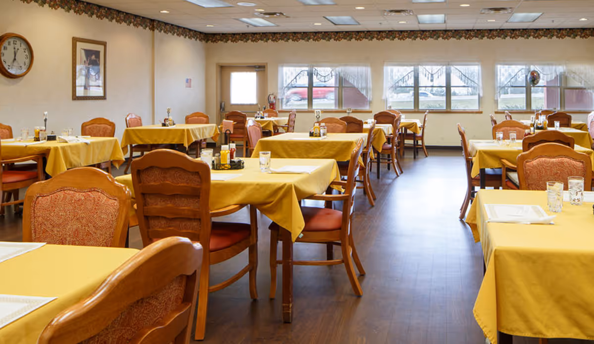 A bright dining room with multiple tables covered in yellow tablecloths and wooden chairs arranged on a hardwood floor.