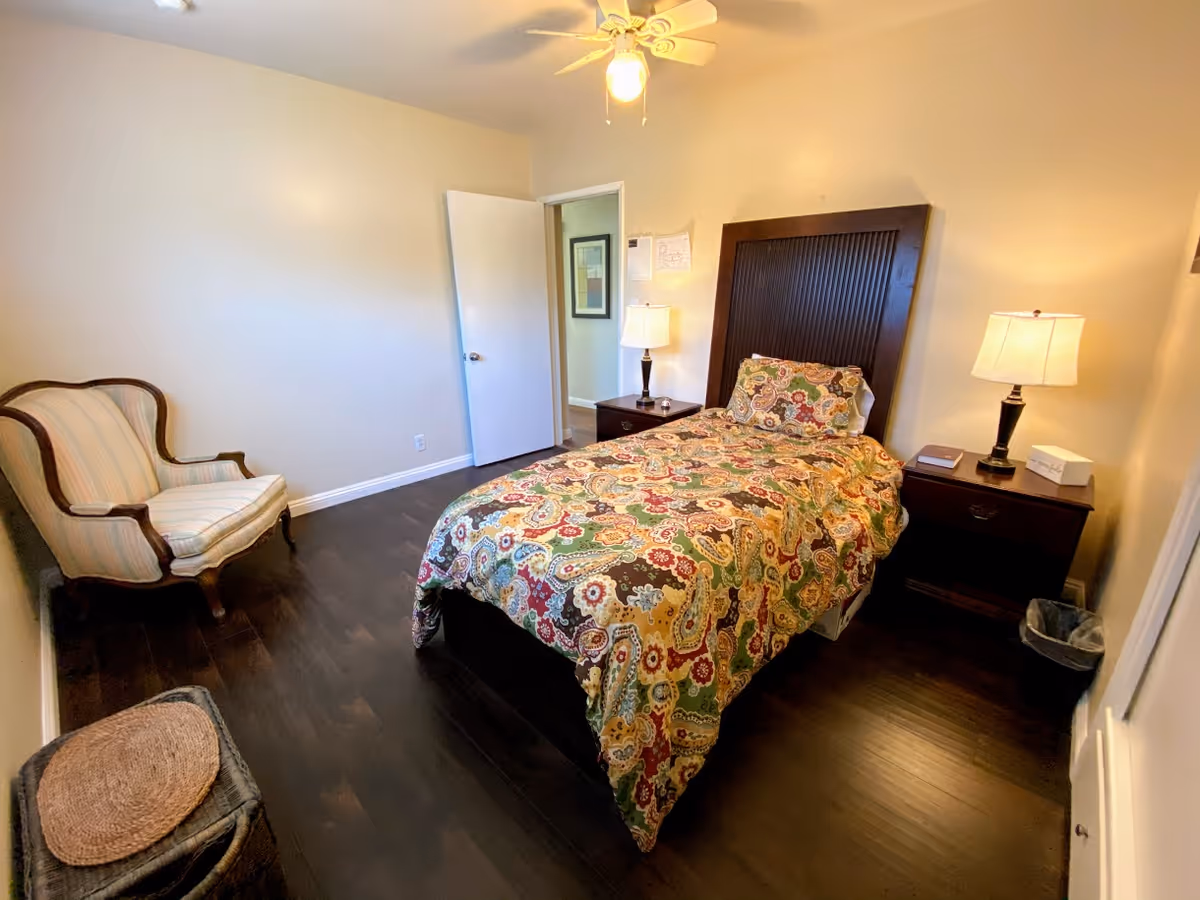 A bedroom with a single bed covered in a colorful patterned bedspread. There are two dark wooden nightstands on either side of the bed, each with a lamp. A cushioned armchair with wooden frame is placed against the left wall. The floor is dark wood, and the walls are painted light beige. A ceiling fan with a light is visible above, and a door is open leading to another room.