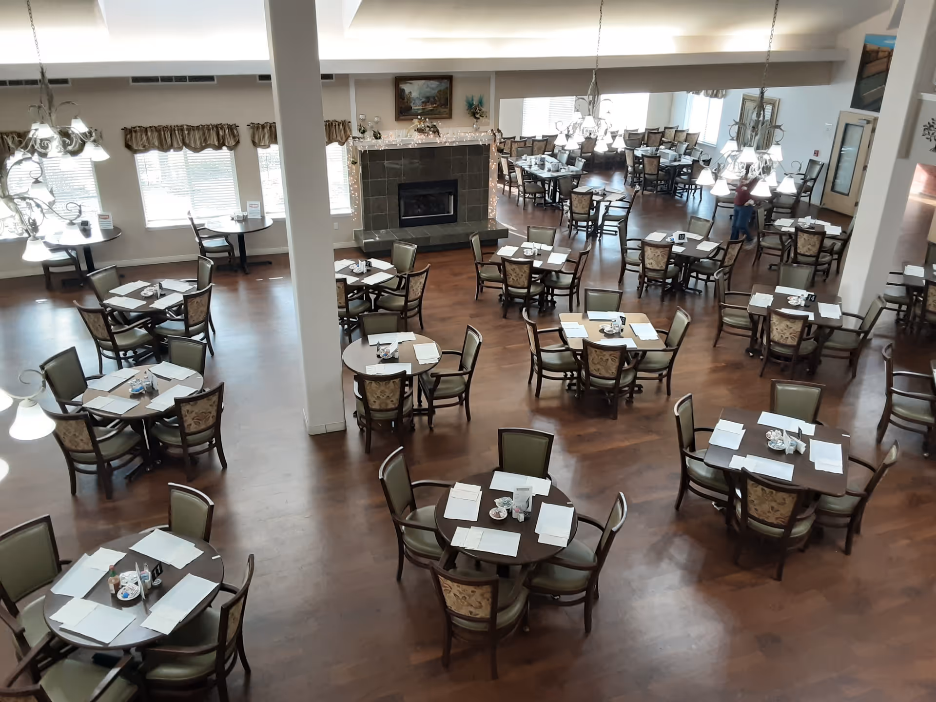 Spacious dining room with multiple round tables and chairs arranged neatly on a wooden floor. Each table is set with placemats, napkins, and condiments. The room features large windows with valance curtains, chandeliers hanging from the ceiling, and a fireplace with decorative items on the mantel.