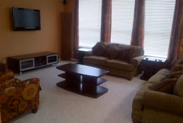 A living room with two brown sofas, a floral patterned armchair, a dark wooden coffee table, a TV mounted on the wall, and a TV stand below it. The room has large windows with blinds and patterned curtains.