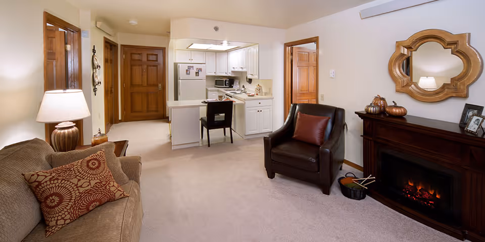 A cozy living room area with a beige sofa adorned with patterned cushions, a dark brown leather armchair with a red pillow, and a wooden electric fireplace with a decorative mirror above it. The room has beige carpet and walls, with a small kitchen area in the background featuring white cabinets, a refrigerator, microwave, and a small dining counter with a chair. Wooden doors and trim add warmth to the space.