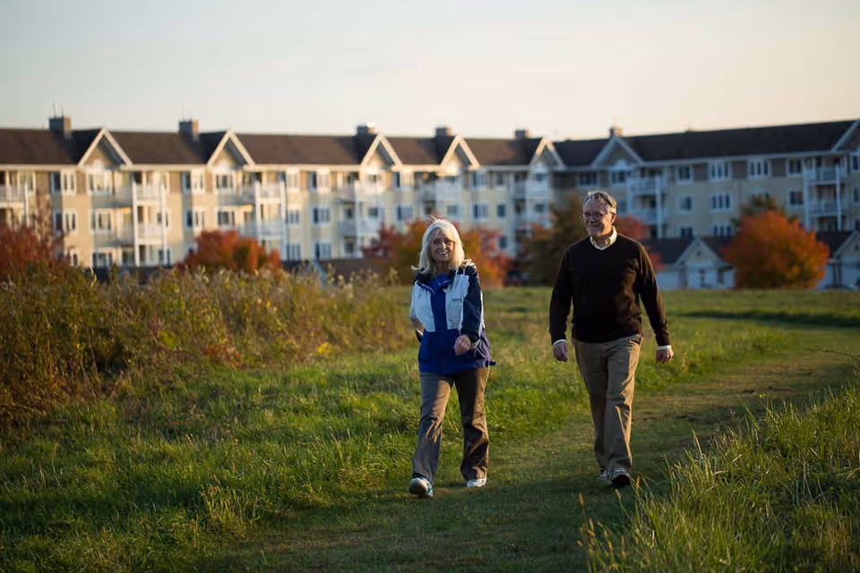 An elderly man and woman walking on a grassy path with a large residential building in the background during late afternoon or early evening.