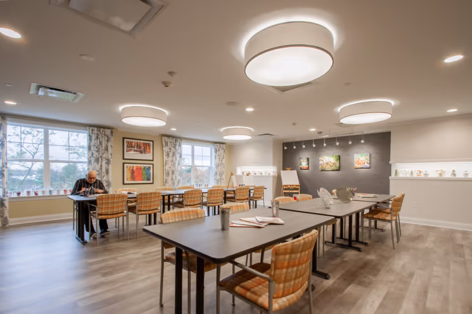 A spacious, well-lit activity or dining room with multiple tables and plaid cushioned chairs arranged neatly. Large windows with patterned curtains allow natural light to fill the room. The walls are decorated with colorful artwork and shelves displaying small decorative items. A man is seated at one of the tables near the windows, engaged in an activity.