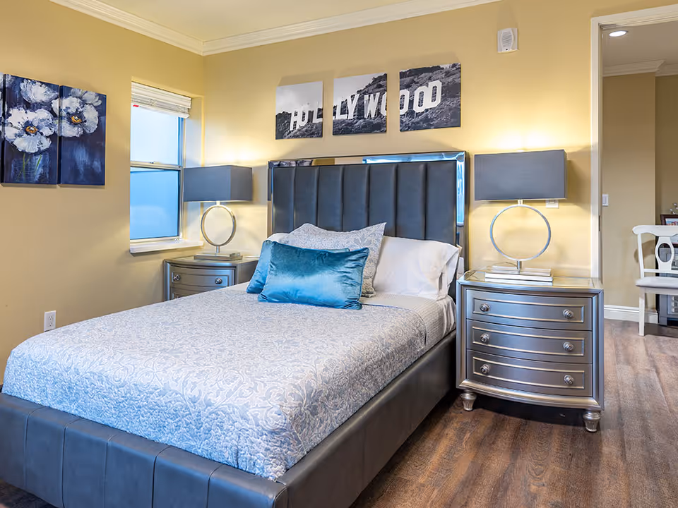 Bright furnished bedroom featuring a bed with a dark upholstered headboard, matching nightstands and lamps, and three-panel Hollywood artwork above the bed.