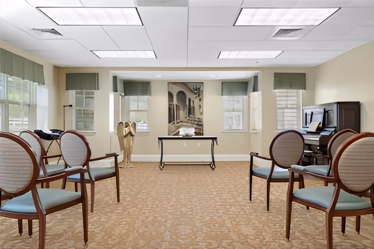 A spacious room with six wooden chairs arranged in two rows facing each other, a piano on the right side, a small table against the far wall, and a wooden angel sculpture near the windows. The room has large windows with green valances, beige walls, and a patterned carpet floor.