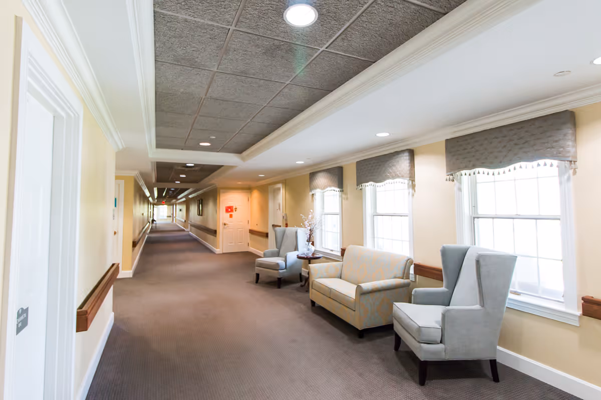 A long, well-lit hallway in a senior living facility with a seating area of armchairs and a loveseat beside windows.