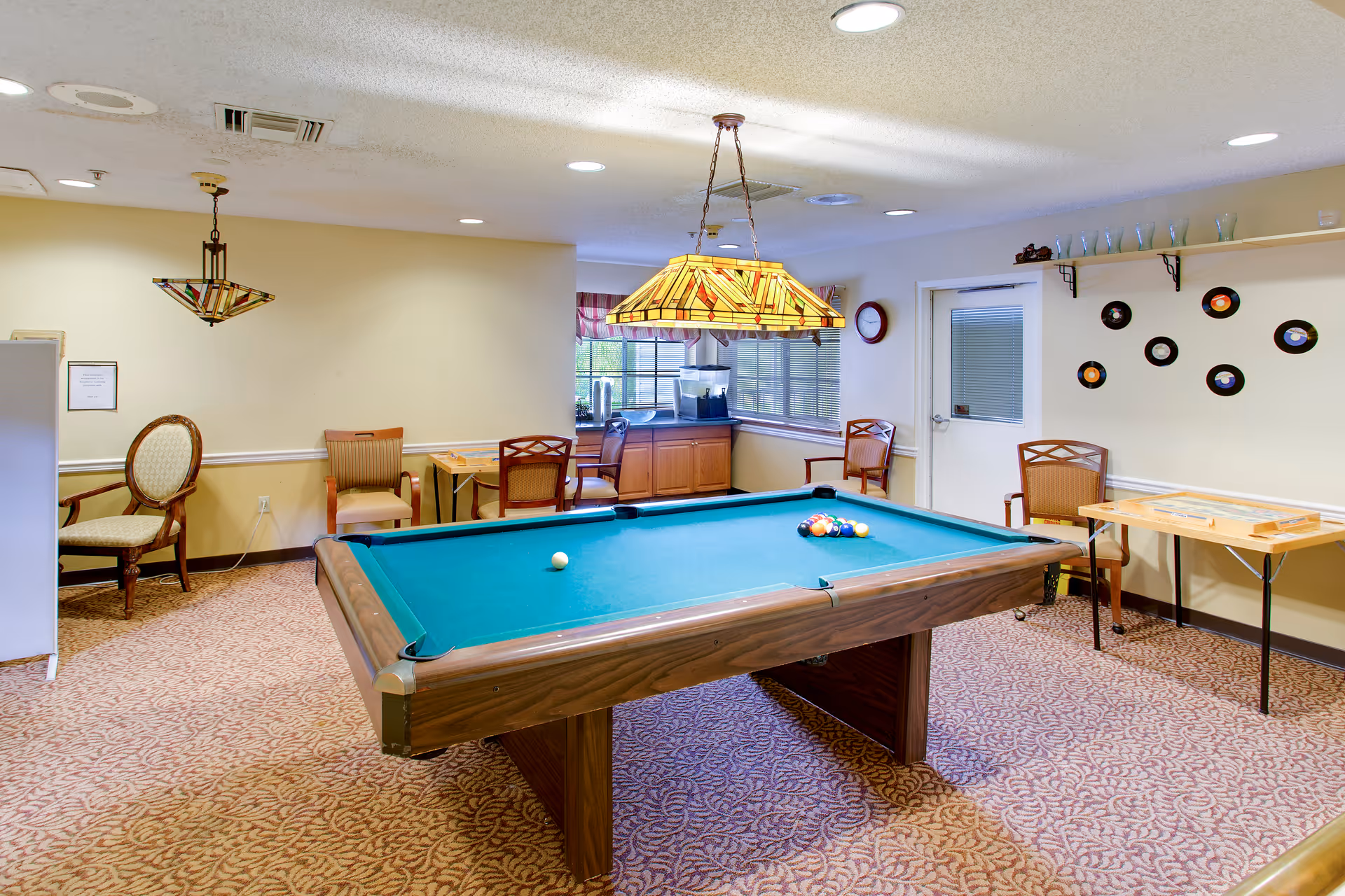 A recreational room with a pool table in the center, surrounded by several chairs and small tables. The room has patterned carpet flooring, a stained glass hanging light fixture above the pool table, and a wall decorated with vinyl records. There is a window with blinds and a door on the right side, and a countertop with a water dispenser in the back corner.