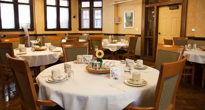 A dining room with round tables covered in white tablecloths, each set with cups, saucers, glasses, and small decorative centerpieces. The room has wooden chairs with green cushioned seats and backs, large windows letting in natural light, and warm yellow wallpaper with a subtle pattern.