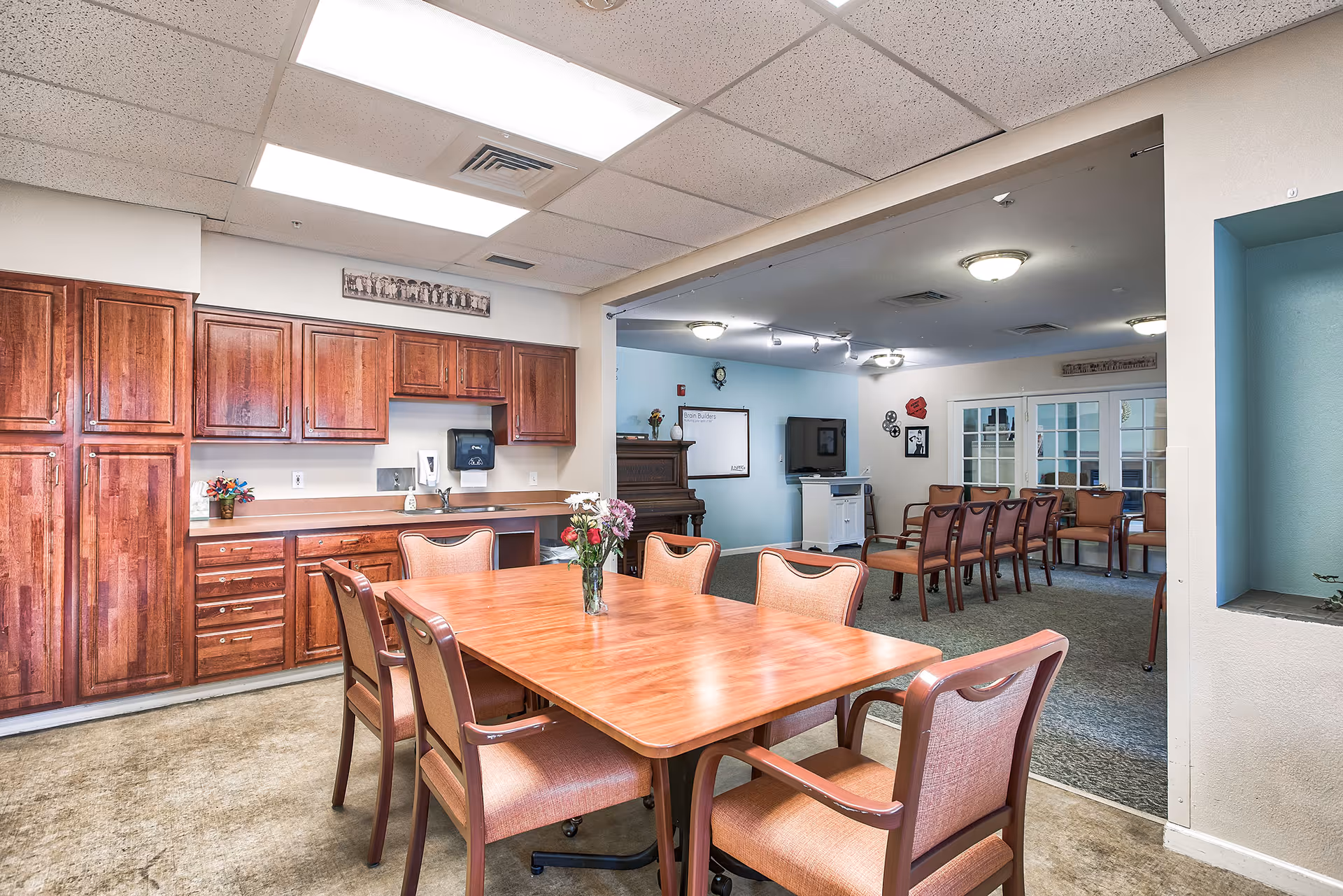 A communal room in a senior living facility featuring a wooden table with six chairs and a vase of flowers in the foreground. Behind the table is a kitchenette with wooden cabinets and a countertop. In the background, there is a lounge area with additional chairs, a piano, a television, and wall decorations.