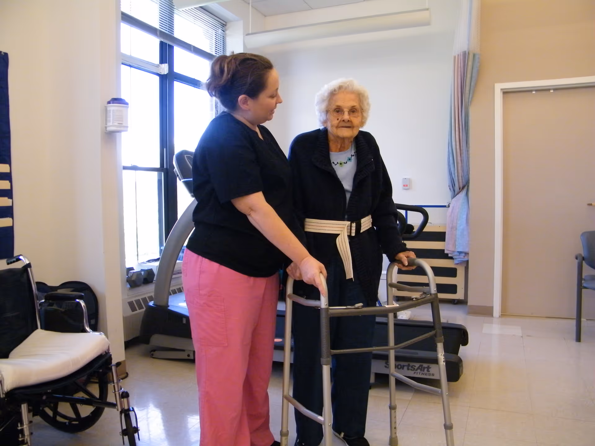 A caregiver assists an elderly woman using a walker in a well-lit room with exercise equipment and a wheelchair nearby.
