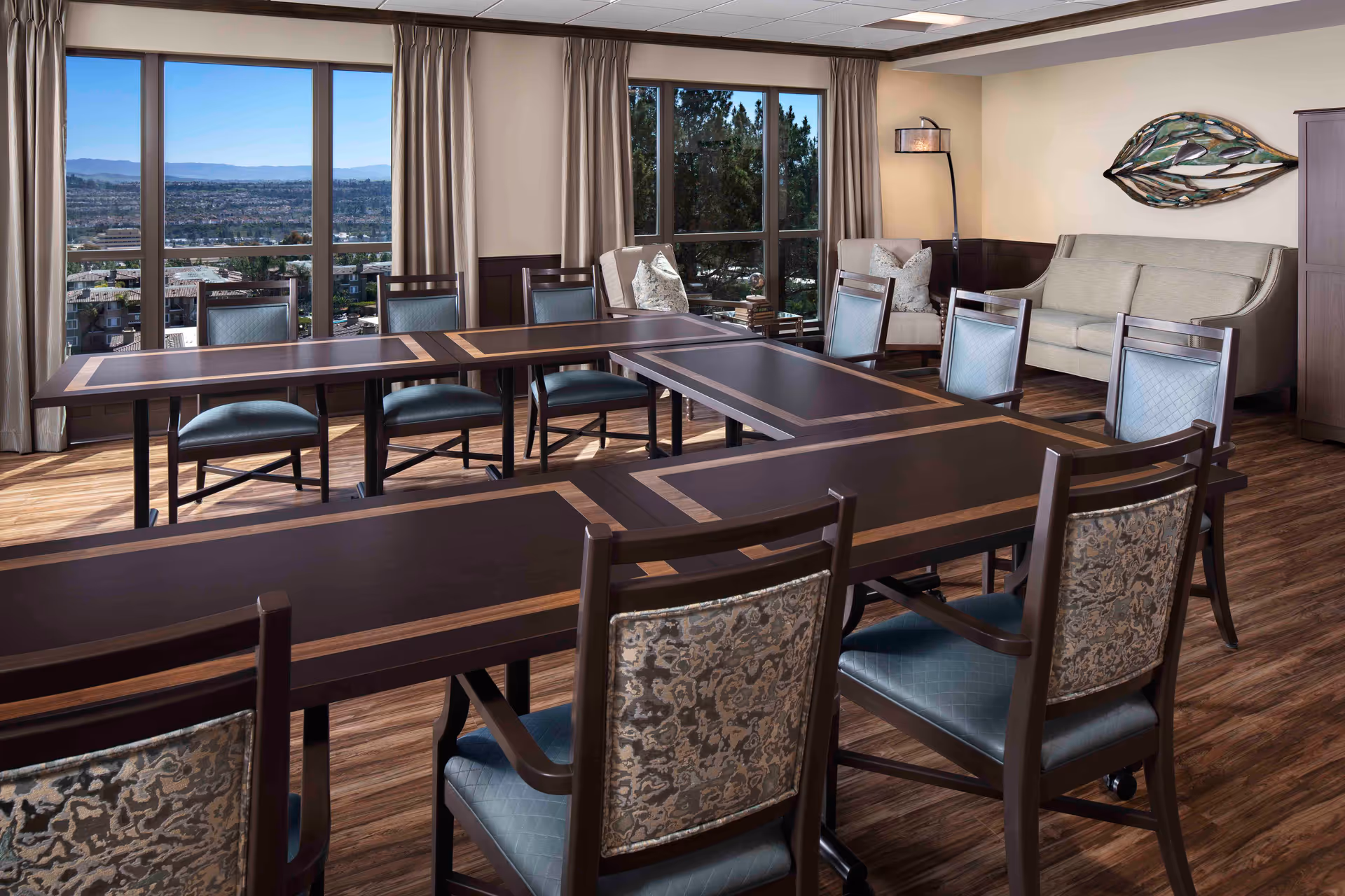 Well-lit communal dining/activity room with U-shaped wooden tables, upholstered chairs, a sofa, and large windows overlooking the city.