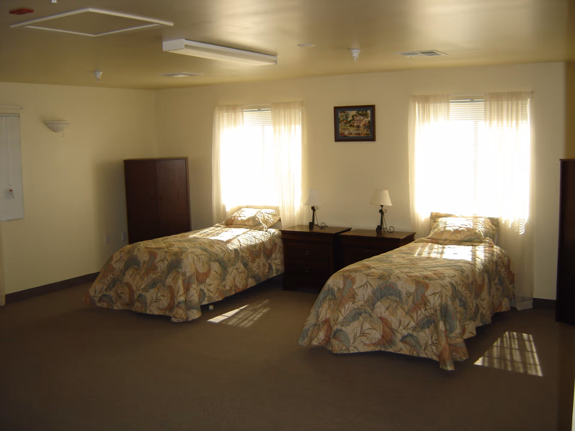 A simple bedroom with two single beds covered in floral patterned bedspreads. Each bed has a pillow and is positioned near a window with sheer curtains allowing sunlight to enter. Between the beds are two wooden nightstands with lamps. There is a wooden cabinet in the corner and a framed picture hanging on the wall between the windows.
