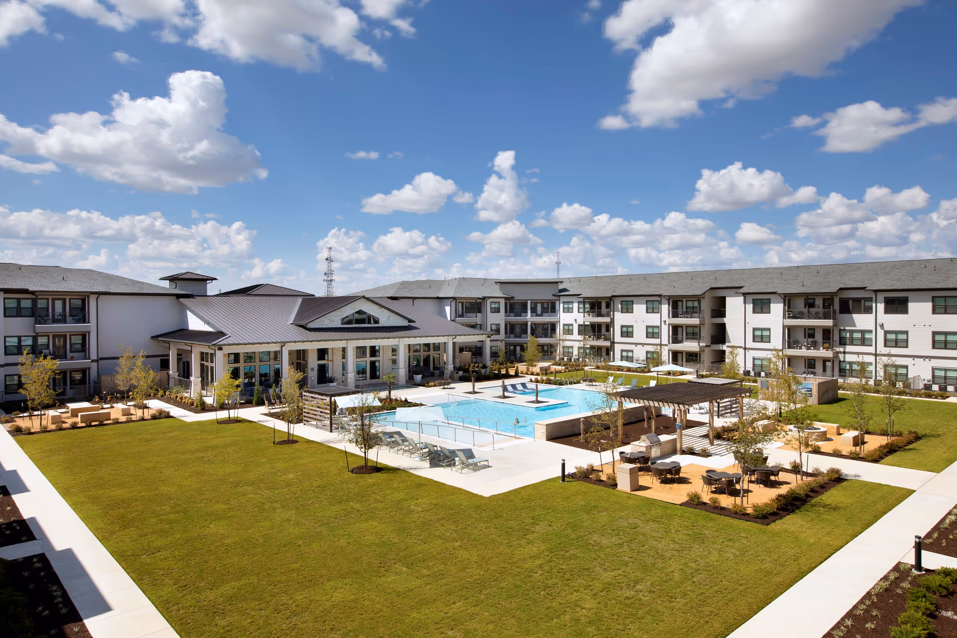 Outdoor view of Solea Keller facility showing a large grassy courtyard with a swimming pool, lounge chairs, shaded seating areas with tables, and surrounding multi-story residential buildings under a partly cloudy blue sky.