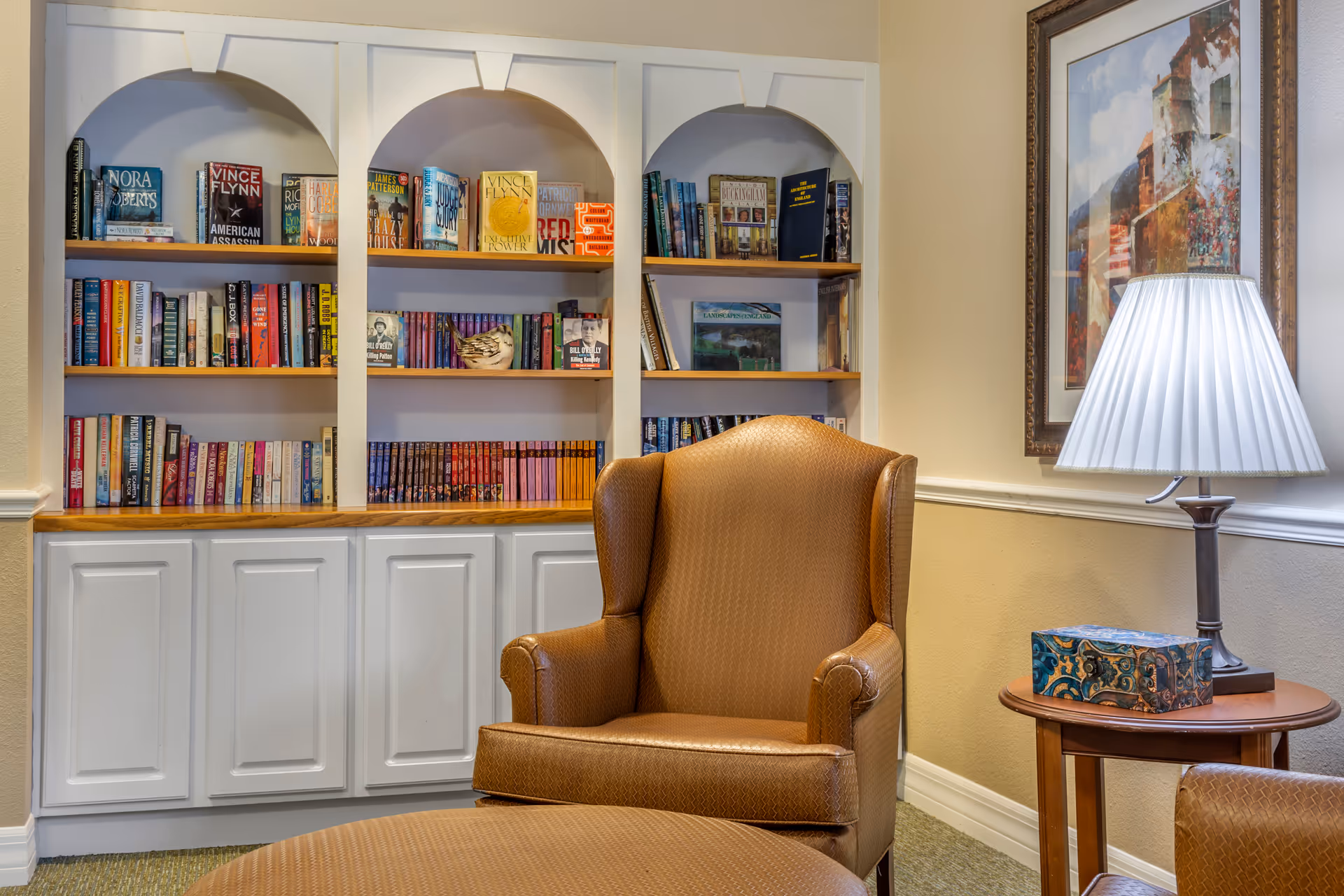 A cozy reading nook featuring a brown upholstered armchair and matching ottoman in front of a built-in white bookshelf filled with books. To the right, there is a wooden side table with a decorative box and a lamp with a white pleated shade. A framed painting hangs on the beige wall behind the lamp.