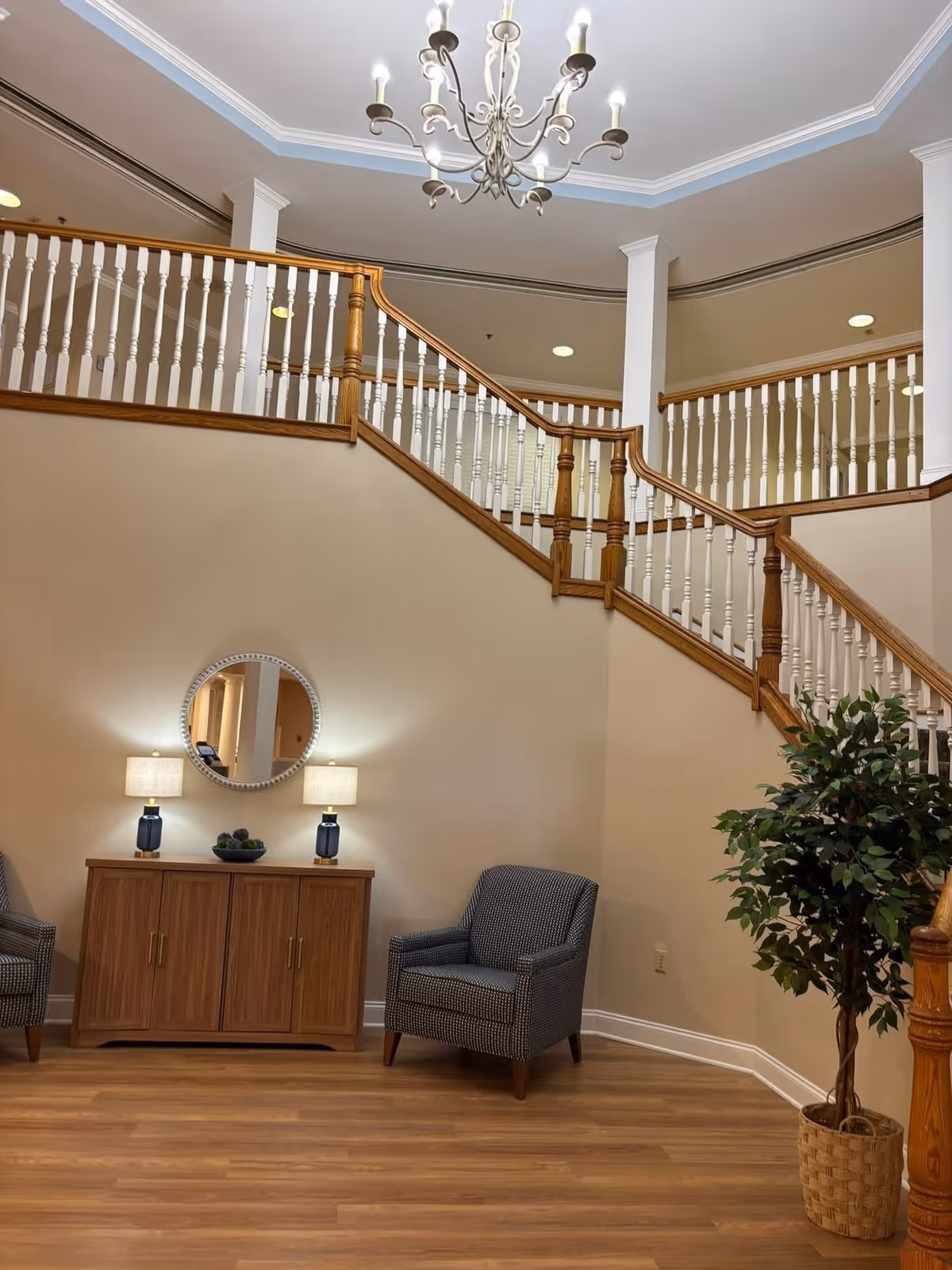 Interior lobby featuring a curved wooden staircase with a chandelier, two armchairs flanking a console table and a potted plant.