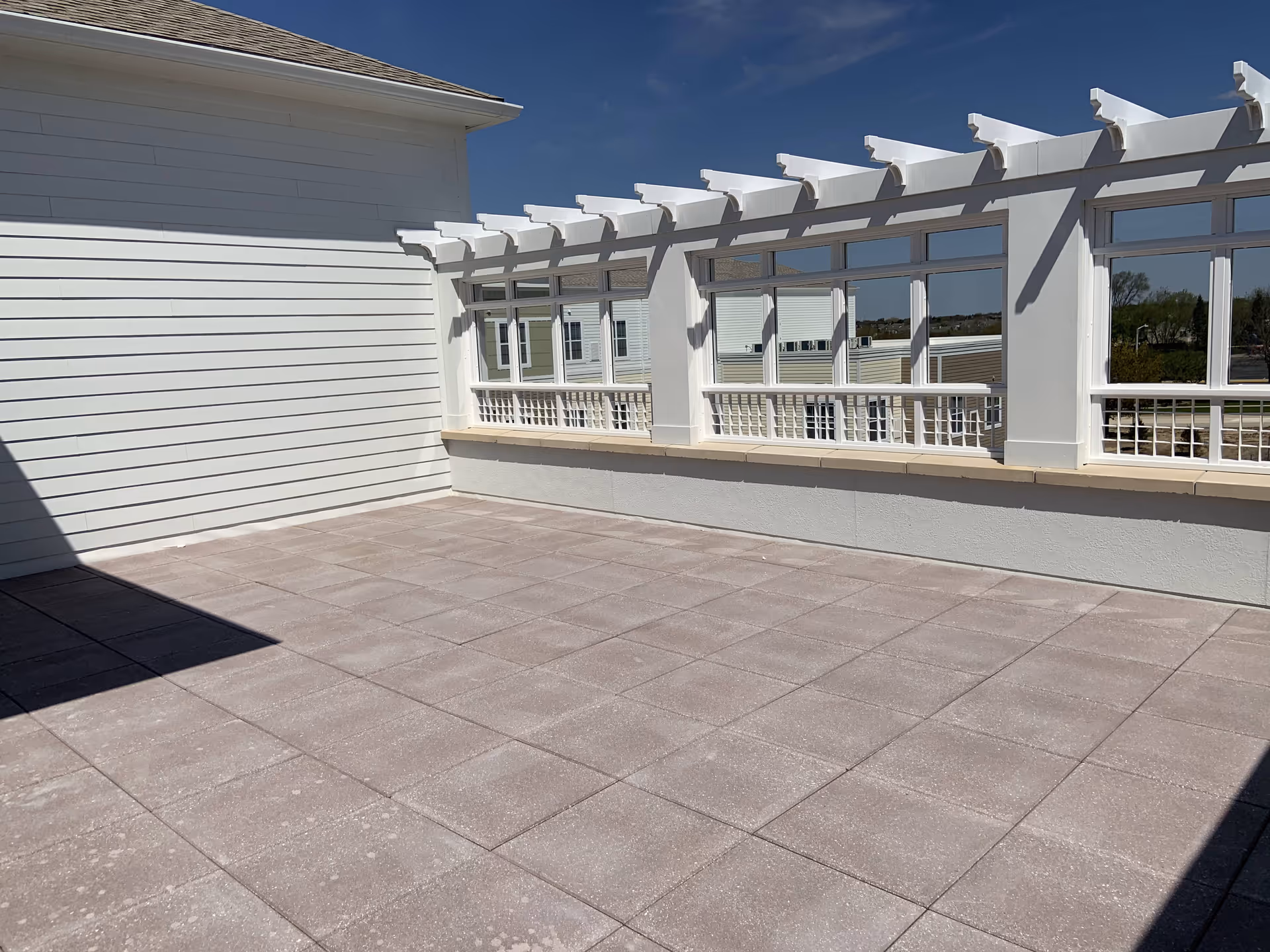 Sunlit outdoor terrace with square tiled floor and a white pergola-style railing along windows.