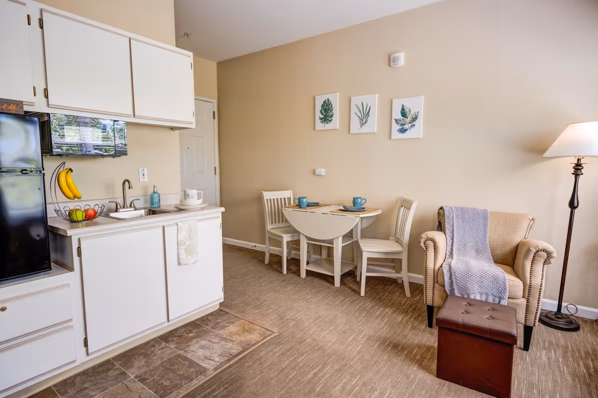 A cozy senior living space featuring a small kitchenette with white cabinets, a black refrigerator, and a sink. Next to the kitchenette is a small dining area with a white drop-leaf table set for two with blue mugs and plates. On the wall above the table are three framed botanical prints. To the right is a beige armchair with a gray knitted throw draped over it, accompanied by a brown ottoman and a tall floor lamp providing warm light.