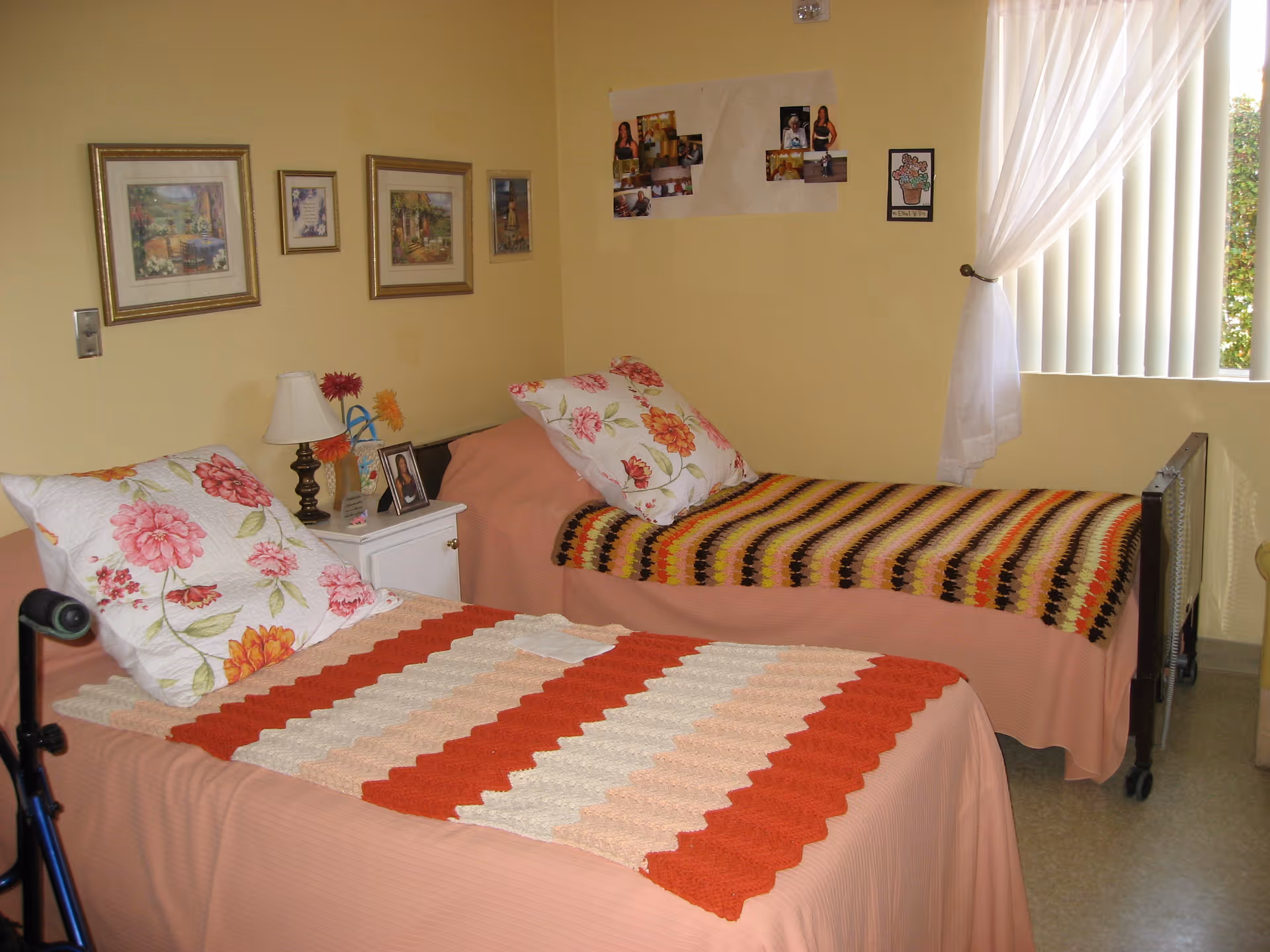 A bedroom with two single beds covered with crocheted blankets and floral pillows. Between the beds is a white nightstand with a lamp, a framed photo, and artificial flowers. The walls are decorated with framed pictures and a collage of photos. A window with vertical blinds and sheer curtains lets in natural light.