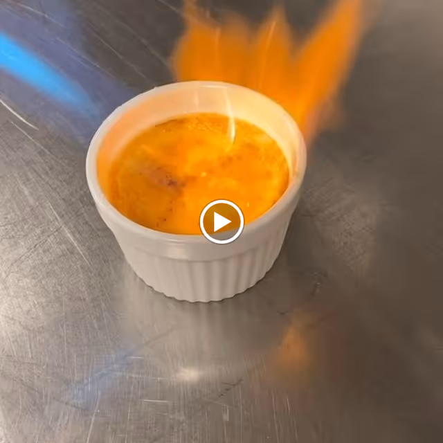 A white ramekin filled with crème brûlée being caramelized with a blue flame torch on a metal surface.