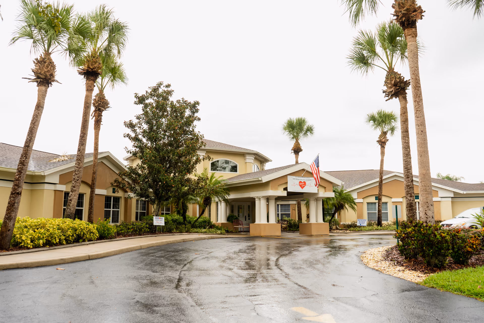 Exterior view of Best Care Senior Living at Brandon facility with a driveway, palm trees, and an American flag near the entrance on a cloudy day.