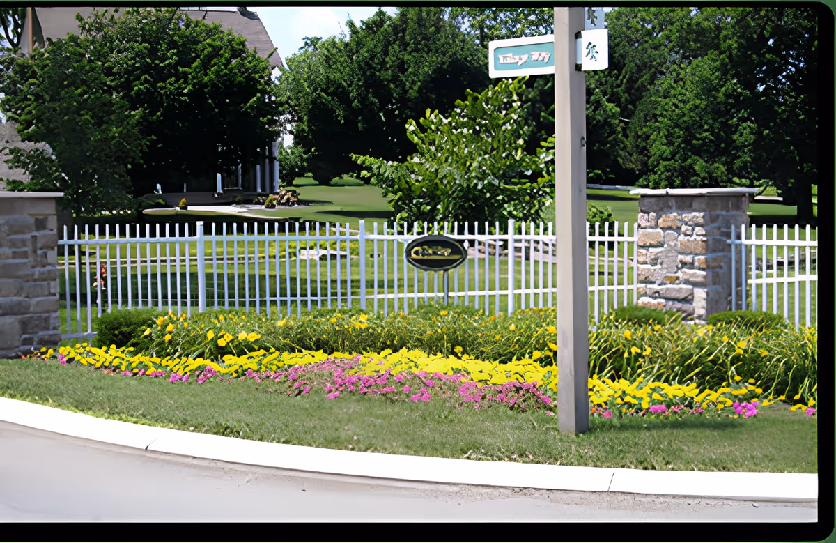 A landscaped garden area with colorful flowers in front of a white picket fence and stone pillars. Behind the fence, there are green lawns and large trees. A wooden post with a small sign is visible near the center-right of the image.