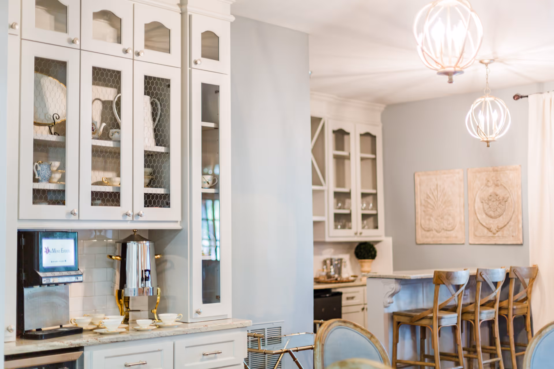 A bright and elegant kitchen area with white cabinetry featuring glass doors displaying dishes and cups. There is a coffee machine and a silver coffee urn on the countertop. In the background, there is a breakfast bar with four wooden stools and two decorative wall hangings. Two modern pendant lights hang from the ceiling, and a window with white curtains is partially visible.