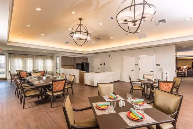 A bright and spacious dining room with several tables set for meals. Each table has colorful plates, glasses, and neatly folded green napkins. The room features modern light fixtures hanging from the ceiling, wood flooring, and large windows with sheer curtains allowing natural light to enter. In the background, there is a kitchen area with a marble countertop and appliances.