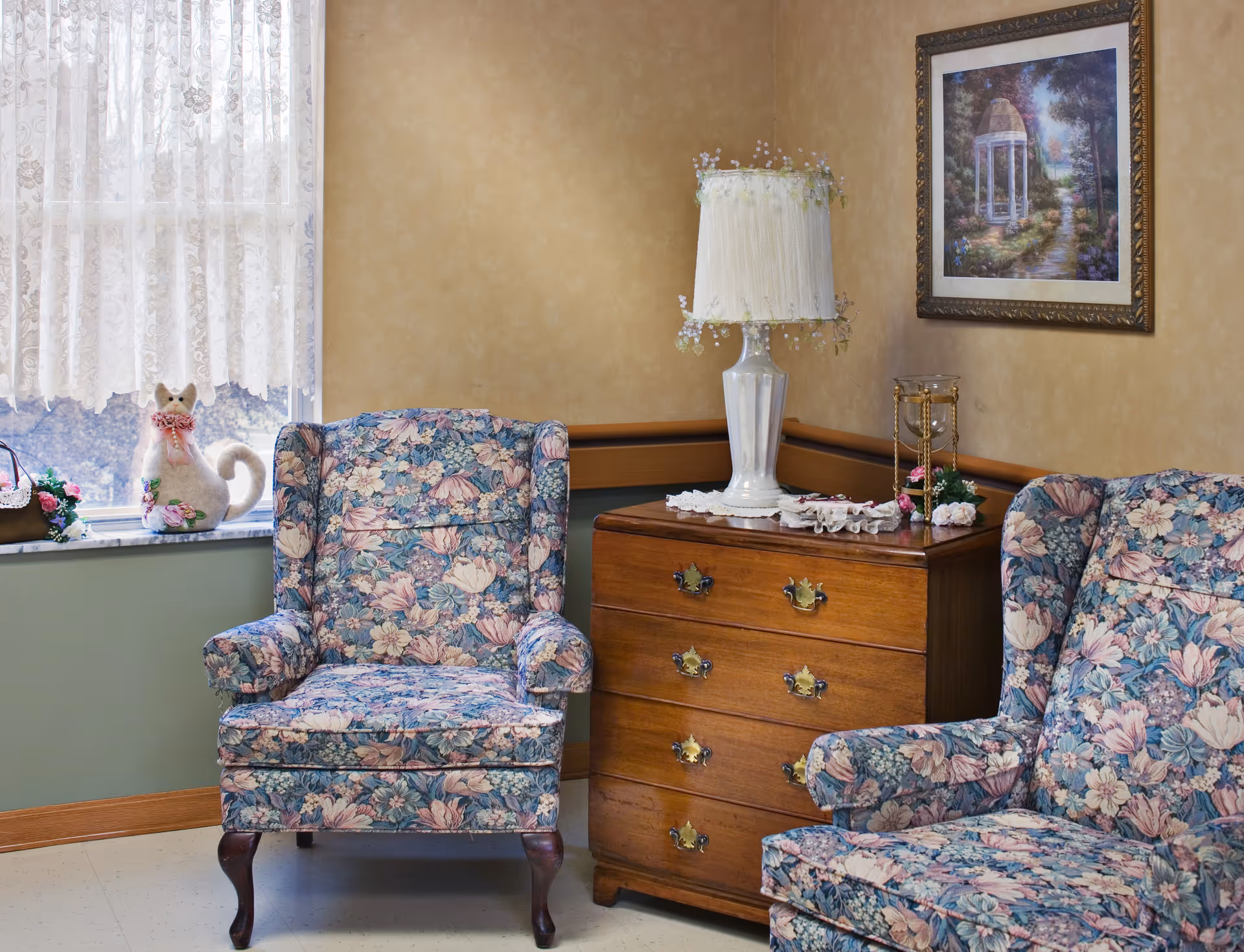 A cozy corner of a room featuring two floral upholstered armchairs, a wooden chest of drawers with brass handles, a decorative lamp with a fringed shade, and a framed painting on the wall. A window with lace curtains and a small cat-shaped decorative item on the windowsill is also visible.