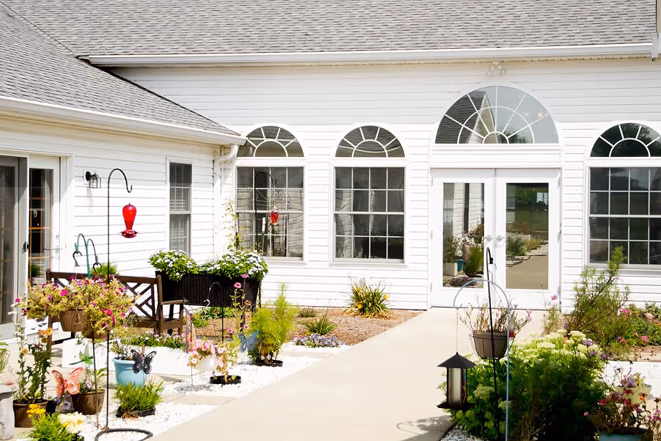Outdoor garden area with various potted plants and flowers along a concrete pathway leading to a white building with large arched windows and glass double doors. There are decorative garden hooks with hanging plants and a red hummingbird feeder. A wooden bench is visible near the building.