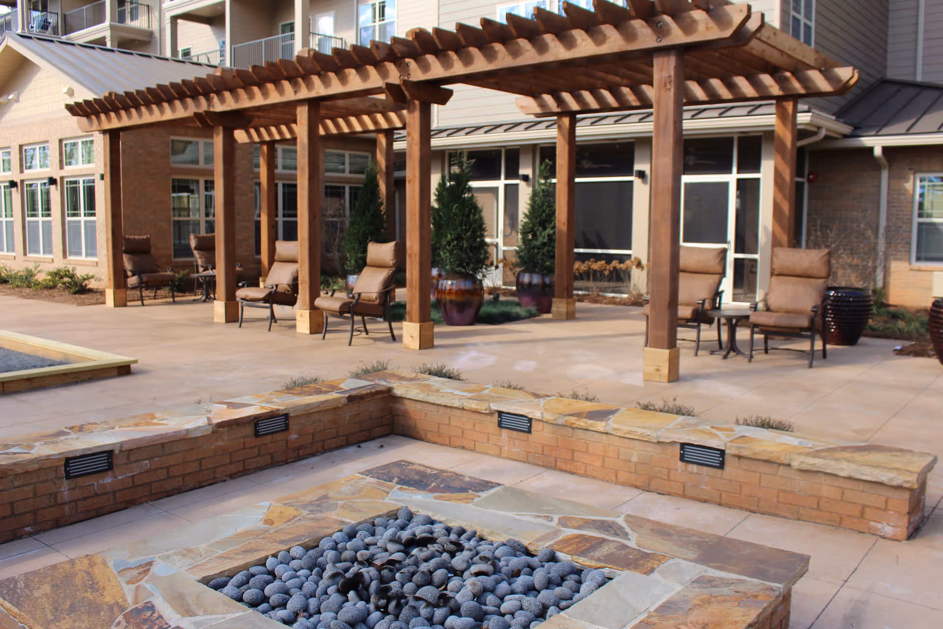 Outdoor patio area at Williams Place Gracious Retirement Living featuring a stone fire pit surrounded by a low brick wall, several cushioned chairs under wooden pergolas, and large potted plants near the building with multiple windows.