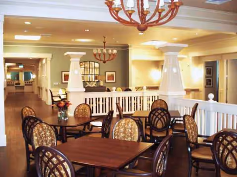 Dining room in a senior living facility with wooden tables, chairs, columns, and chandeliers.