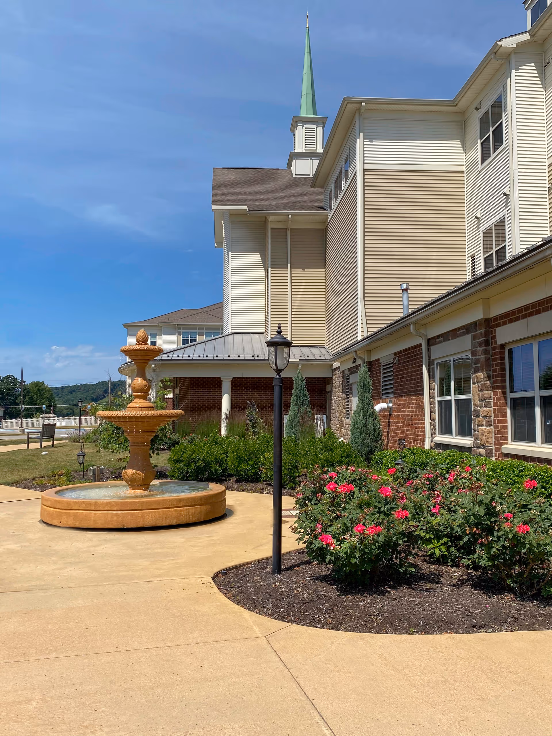 Outdoor view of a senior living facility with a beige and brick exterior, a three-tiered water fountain, a black lamp post, and landscaped flower beds with pink flowers under a clear blue sky.