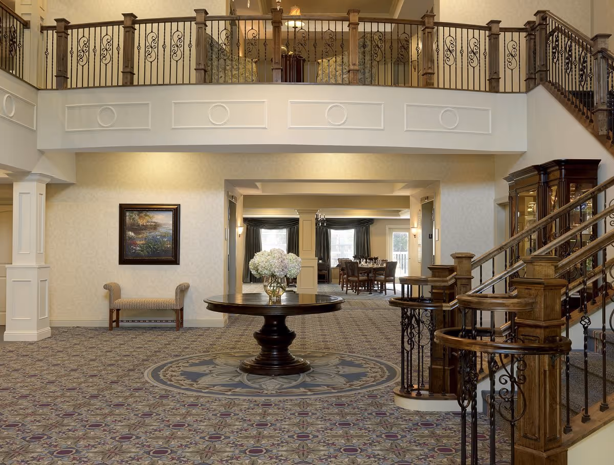 Elegant two-story lobby with a central round table holding a floral arrangement, decorative carpeting, staircase and wrought-iron balcony railing.