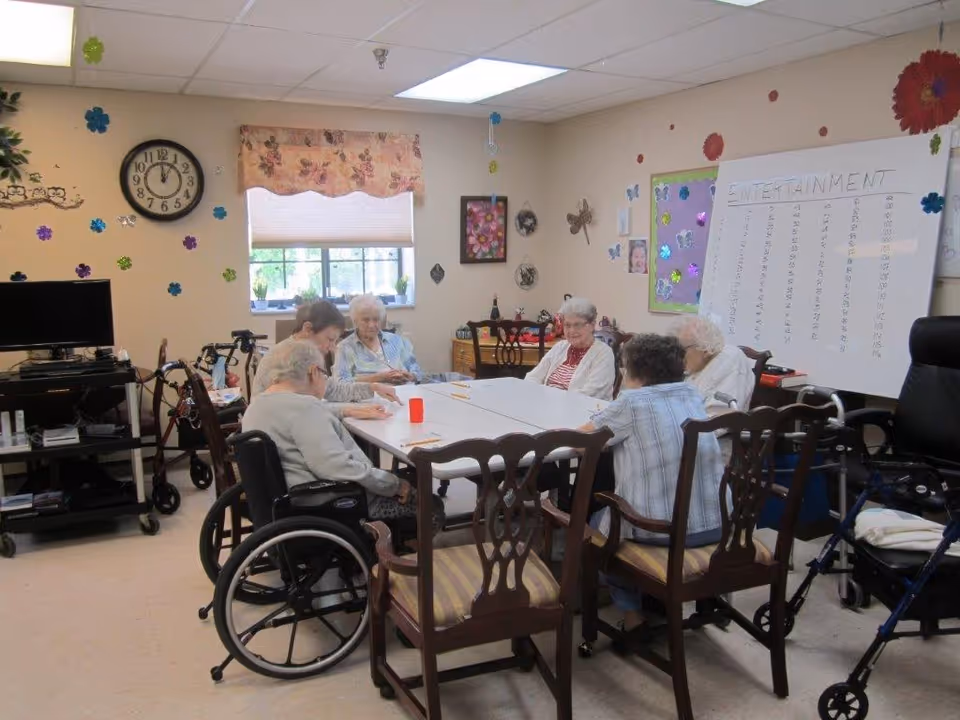 A group of elderly individuals sitting around a rectangular table in a decorated room with floral wall decals and a clock showing 11:55. Some are seated in wheelchairs and others in wooden chairs. There is a whiteboard with the word 'ENTERTAINMENT' written on it and a television on a stand to the left.