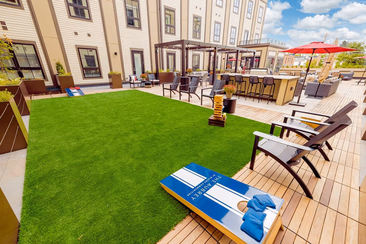 Outdoor patio area at Upside Columbus featuring artificial grass with a cornhole game set up, Adirondack chairs, a large Jenga game, bar seating with stools under pergolas, and red umbrellas providing shade over seating areas. The building exterior with windows and doors is visible in the background under a partly cloudy sky.