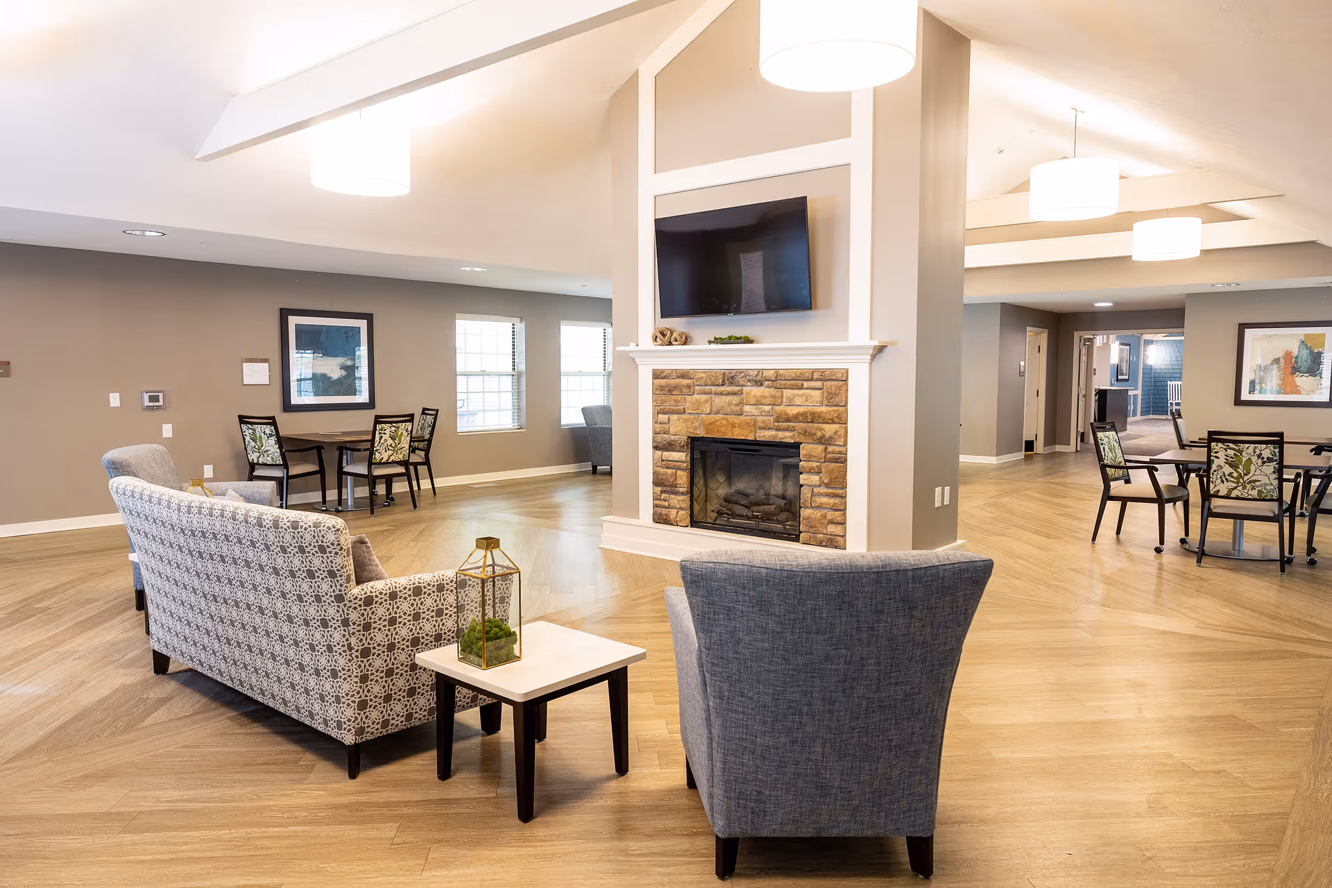 A spacious senior living common area with a stone fireplace and a mounted flat-screen TV. The room features comfortable seating including a patterned sofa and a gray armchair, small tables, and multiple dining tables with chairs. The walls are painted light gray and decorated with framed artwork. The floor is wood with a herringbone pattern, and the ceiling has modern light fixtures.