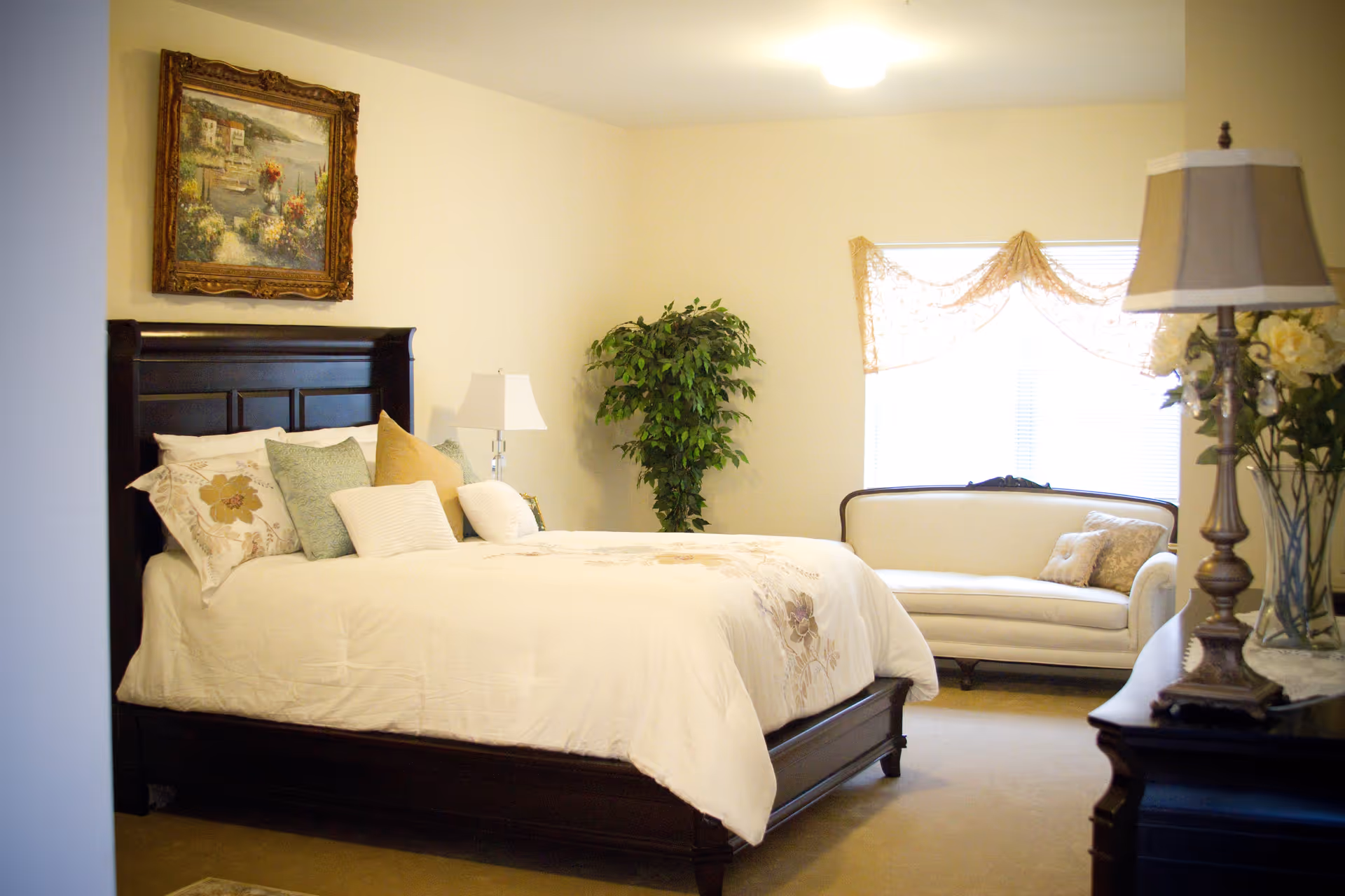 A cozy bedroom with a dark wooden bed frame and a white comforter adorned with floral patterns and multiple pillows. There is a white lamp on a nightstand beside the bed, a green potted plant in the corner, a white upholstered loveseat near a window with sheer curtains, and a wooden dresser with a decorative lamp and flowers on it. A framed painting hangs on the wall above the bed.