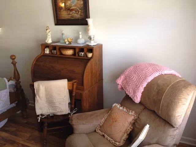 A cozy corner of a room featuring a beige recliner chair with a decorative pillow and a pink knitted throw draped over the back. Next to the chair is a wooden roll-top desk with various decorative items on top, including figurines and a lamp. A wooden chair with a beige cloth draped over the back is positioned in front of the desk. The room has wooden flooring and a framed painting hanging on the wall above the desk.