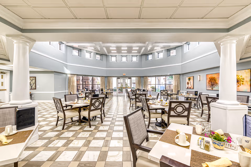 Bright, spacious dining room with a checkered floor, multiple set tables, tall columns and a high skylit ceiling.
