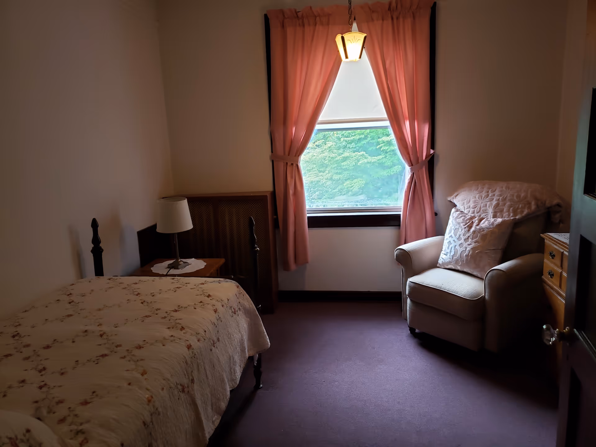 Small bedroom with a single bed and nightstand, an upholstered armchair by a window with pink curtains, and a hanging light.